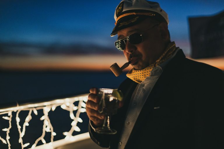 A man in a captain's hat smokes a cigar and holds a cocktail in his hand while standing on a boat.